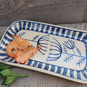 Może przedstawiać: Biała ceramiczna tacka do serwowania z niebieskimi i białymi paskami wokół krawędzi. Taca ma niebieski wzór kwiatowy i dwie gotowane krewetki na górze.