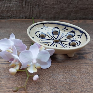 Może przedstawiać: Kremowa ceramiczna mydelniczka z niebieskim kwiatowym wzorem i trzema małymi nóżkami. Owalna miska ma otwory drenażowe i jest ozdobiona niebieskimi liniami i kropkami. Gałązka białych i fioletowych orchidei znajduje się na pierwszym planie.