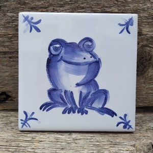 May include: A blue and white ceramic tile with a hand-painted design of a smiling frog. The tile has a white background with blue floral accents in the corners.
