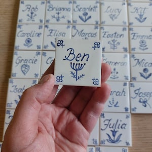 Könnte beinhalten: Eine Hand hält eine weiße Keramikfliese mit dem Namen "Ben" in blauer Schrift, umgeben von floralen und geometrischen Mustern. Weitere Fliesen mit Namen wie "Mae" und "Julia" sind im Hintergrund zu sehen, alle mit ähnlichen blauen Designs.