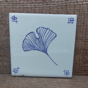 Op de afbeelding: Vierkante witte keramische tegel met een blauw ginkgo blad ontwerp in het midden. De tegel heeft in elke hoek een blauw bloemenontwerp. Deze tegel is waarschijnlijk bedoeld voor gebruik in een keuken of badkamer, ongeveer 15 cm x 15 cm.