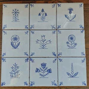 May include: Nine square ceramic tiles arranged in a 3x3 grid. Each white tile features a hand-drawn blue floral design. The flowers vary in style, including thistle, bell-shaped, and dandelion-like motifs. Small blue corner accents complete the design.