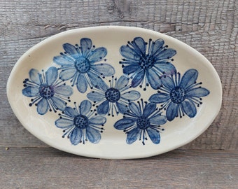 May include: An oval ceramic serving dish with a white background and a blue floral design. The dish features seven stylised blue flowers with white centres.
