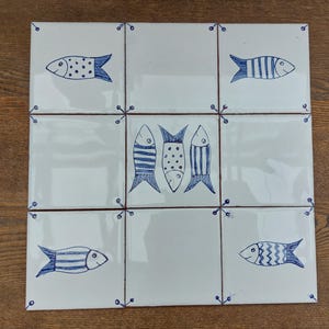 Puede incluir: Nueve baldosas de cerámica blancas dispuestas en un cuadrado, cada una con un diseño de pez azul dibujado a mano. Los peces varían en patrón, algunos muestran rayas o puntos. Las baldosas están delineadas con un borde rojo fino y detalles decorativos azules en las esquinas.
