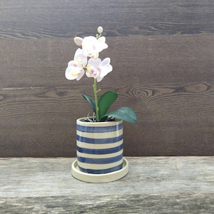 Op de afbeelding: Een keramische plantenpot met blauwe en witte horizontale strepen, met een kunstorchidee met witte en roze bloemen en groene bladeren. De pot staat op een bijpassende schotel. De achtergrond is een donker houten paneel.