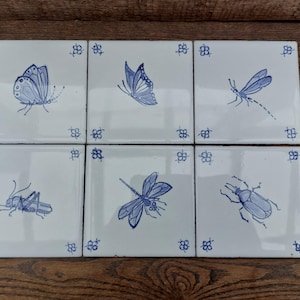 May include: Six white ceramic tiles with blue insect illustrations, including butterflies, dragonflies, grasshoppers, and beetles. Each tile has a blue floral corner design. The tiles are arranged on a wooden surface.