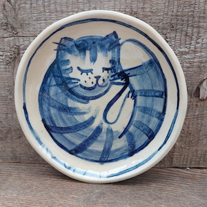 Peut inclure: Une assiette en céramique blanche avec un motif peint à la main d'un chat enroulé. Le chat est représenté avec un motif rayé bleu et blanc et un visage souriant.