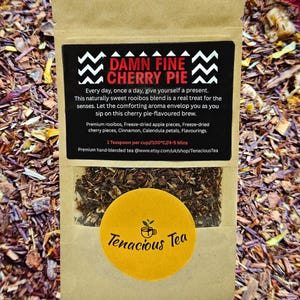 May include: A brown paper bag of "Damn Fine Cherry Pie" tea. The black label includes text about the tea's ingredients and brewing instructions. A yellow sticker with the words "Tenacious Tea" is on the front.