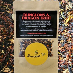 May include: A brown paper bag of loose leaf tea labeled "DUNGEONS & DRAGON FRUIT." The label includes a description of the tea's ingredients and brewing instructions. A yellow sticker with the words "Tenacious Tea" is on the bag.