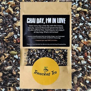 Könnte beinhalten: Eine Packung Tenacious Tea Chai-Mischung. Die Verpackung hat ein schwarzes Etikett mit weißem Text, auf dem "CHAI DAY, I'M IN LOVE" steht. Ein gelber runder Aufkleber zeigt den Markennamen. Die Teemischung enthält Schwarztee, Ingwer, Kokosnuss, Zimt und Kardamom.