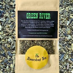 Könnte beinhalten: Eine Packung entkoffeinierten Sencha-Grüntees mit der Aufschrift "GREEN RIVER". Die Verpackung enthält ein schwarzes Etikett mit weißem Text, der die Eigenschaften des Tees und die Brühhinweise beschreibt. Ein gelber runder Aufkleber mit dem Logo "Tenacious Tea" ist ebenfalls sichtbar.