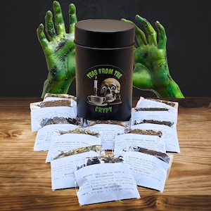 Könnte beinhalten: Eine schwarze Teedose aus Metall mit einem Totenkopf- und Kerzengrafik, beschriftet mit "Teas From The Crypt", umgeben von Teepäckchen. Das Bild enthält grüne, zombieartige Hände im Hintergrund. Die Teepäckchen haben Brühhinweise.