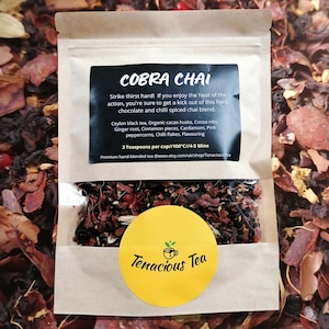 Può includere: Un sacchetto di carta marrone pieno di tè sfuso. L'etichetta nera recita "COBRA CHAI" ed elenca ingredienti come tè nero di Ceylon e fave di cacao. Un adesivo giallo con "Tenacious Tea" è sulla busta. Il tè è fatto a mano.