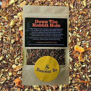 May include: A brown paper bag of loose leaf tea with a black label that reads "Down The Rabbit Hole." The label lists ingredients like rooibos, carrot pieces, and spices. A yellow sticker with "Tenacious Tea" is on the bag.