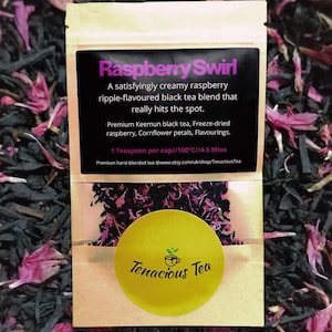 May include: A small, tan tea bag with a black label that reads "Raspberry Swirl." The bag contains a blend of black tea, freeze-dried raspberry, and cornflower petals. A yellow sticker with the words "Tenacious Tea" is on the bag.