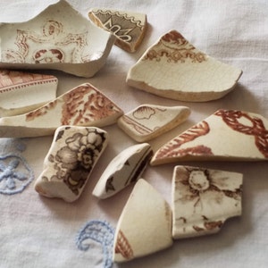 May include: A collection of broken pieces of white ceramic with brown floral patterns. The pieces are arranged on a white fabric surface with blue embroidery.
