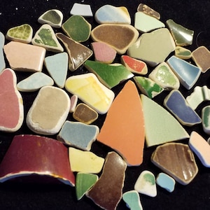 May include: A collection of colorful, broken ceramic tiles in various shapes and sizes. The tiles are arranged in a loose, organic pattern on a black background. The tiles are mostly green, brown, blue, red, and yellow.