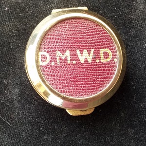 Può includere: Un compatto dorato con un centro strutturato bordeaux. Il centro presenta le iniziali dorate "D.M.W.D.". Il compatto è rotondo e ha un'apertura a cerniera. Lo sfondo è un tessuto scuro e strutturato.