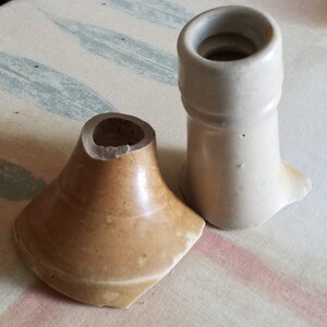May include: Two ceramic bottle stoppers, one is a light brown color and the other is a light gray color. Both stoppers have a round opening at the top and a flared base.