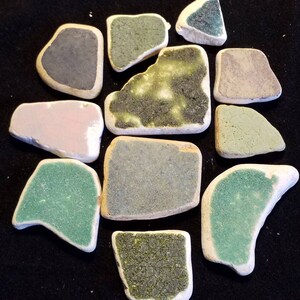 May include: A collection of 11 ceramic tiles in various shades of green, blue, pink, and gray. The tiles are irregularly shaped and have a rustic, handmade appearance.