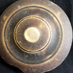 Puede incluir: Un plato de cerámica marrón con un borde elevado y un diseño circular en el centro. El plato tiene una forma ligeramente ovalada y una superficie texturizada.