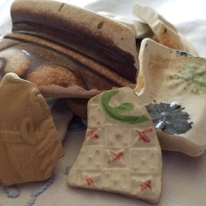 May include: A collection of broken ceramic pieces with various patterns and colors, including brown, white, green, and blue. The pieces are arranged on a white fabric surface.