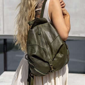 May include: An olive green leather backpack featuring a vertical zippered front pocket, a top handle, and adjustable shoulder straps. The bag has a rounded silhouette with visible stitching details and antique brass-toned metal hardware accents.