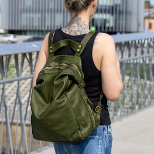 Puede incluir: Una mochila de cuero verde oliva con un diseño suave y estructurado, que presenta múltiples cremalleras metálicas doradas, correas ajustables para los hombros y costuras reforzadas en los puntos de unión del asa superior con remaches de latón visibles.