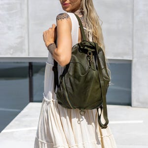 May include: An olive green leather backpack featuring a soft, slouchy silhouette, a top handle, adjustable shoulder straps, and a central brass-toned metal clasp closure. The bag includes visible stitched paneling and a side zipper detail.