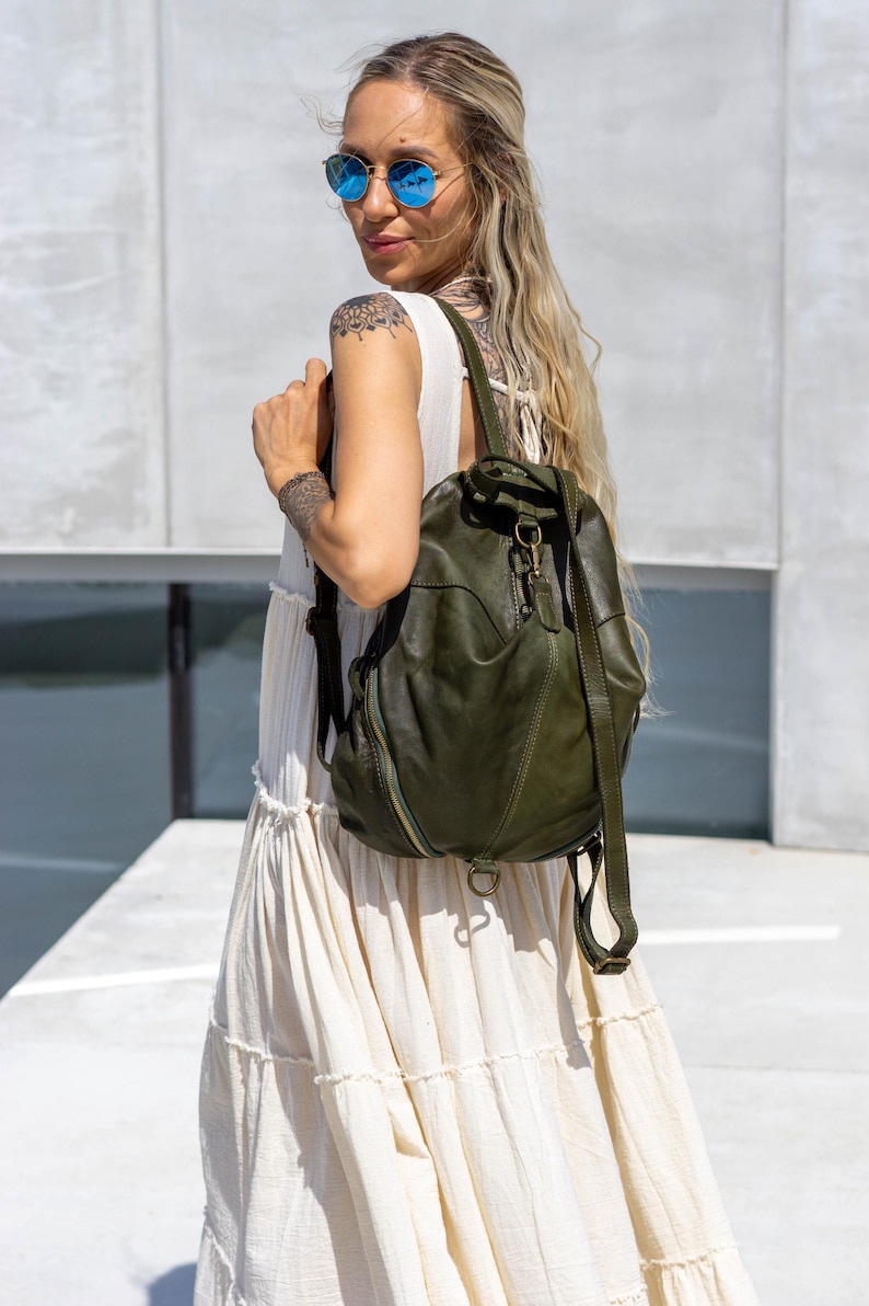 May include: An olive green leather backpack featuring a soft, slouchy silhouette, a top handle, adjustable shoulder straps, and a central brass-toned metal clasp closure. The bag includes visible stitched paneling and a side zipper detail.