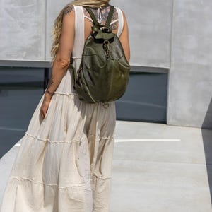 May include: An olive green leather backpack featuring a rounded silhouette, a top flap with a metal clasp closure, and side zips. The bag has adjustable shoulder straps and a structured design, showcasing a smooth texture and visible stitching details.