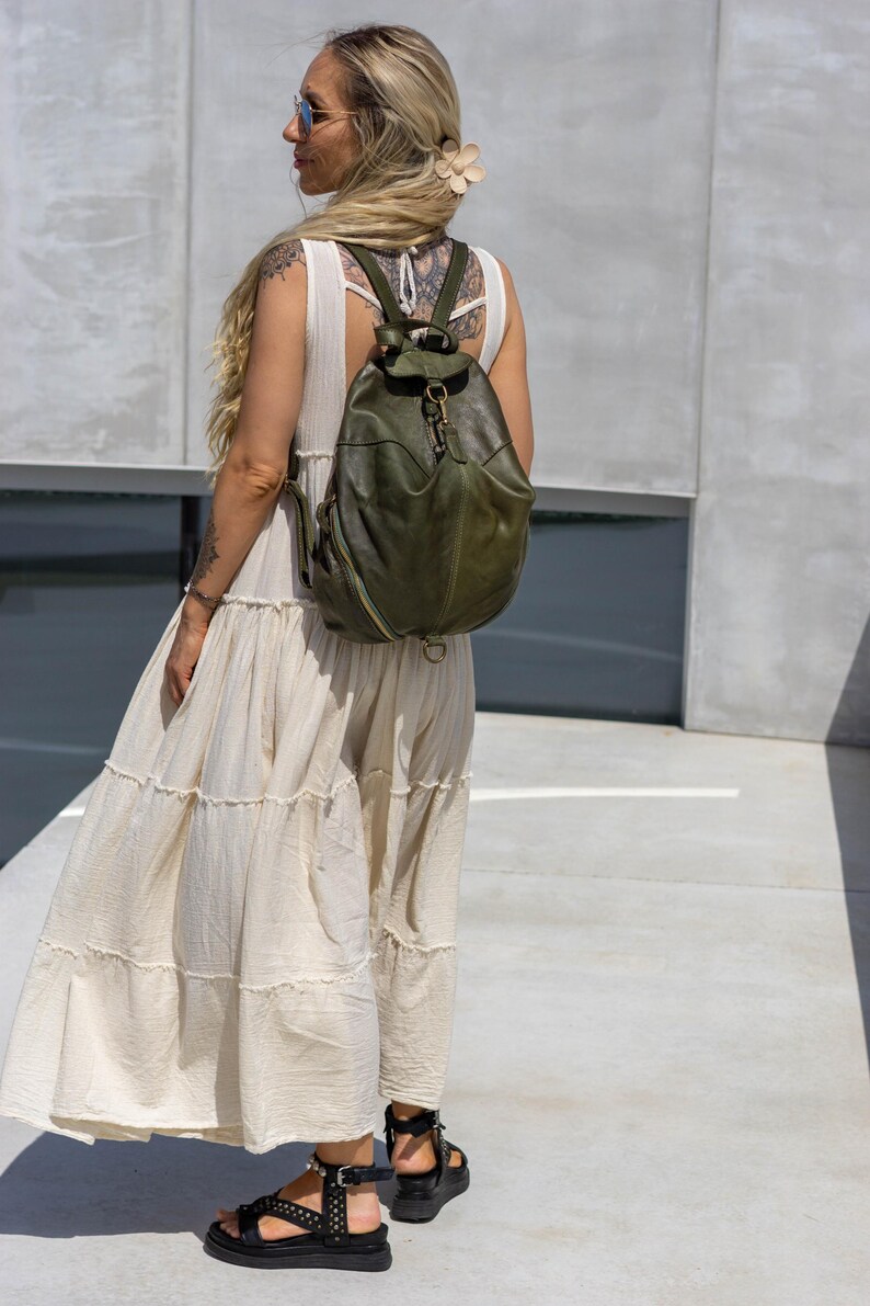 May include: An olive green leather backpack featuring a rounded silhouette, a top flap with a metal clasp closure, and side zips. The bag has adjustable shoulder straps and a structured design, showcasing a smooth texture and visible stitching details.