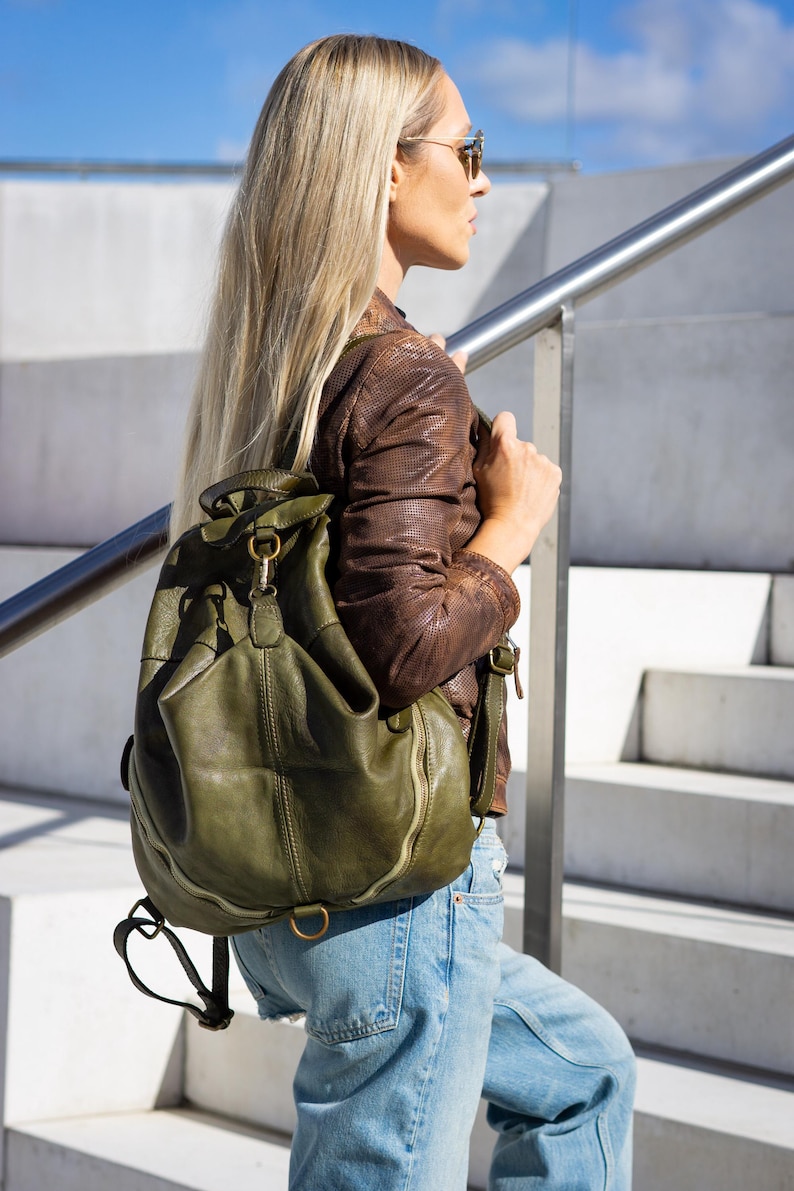 May include: An olive green leather backpack featuring a slouchy silhouette, a top handle, adjustable shoulder straps, and a front flap closure secured with a metal clasp. The bag includes side zips and a bottom metal D-ring for added utility.