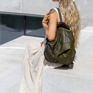 May include: An olive green leather backpack with a rounded silhouette, featuring a central vertical zip, a top flap closure, and adjustable shoulder straps. The bag has a smooth texture and antique brass-toned metal hardware accents.