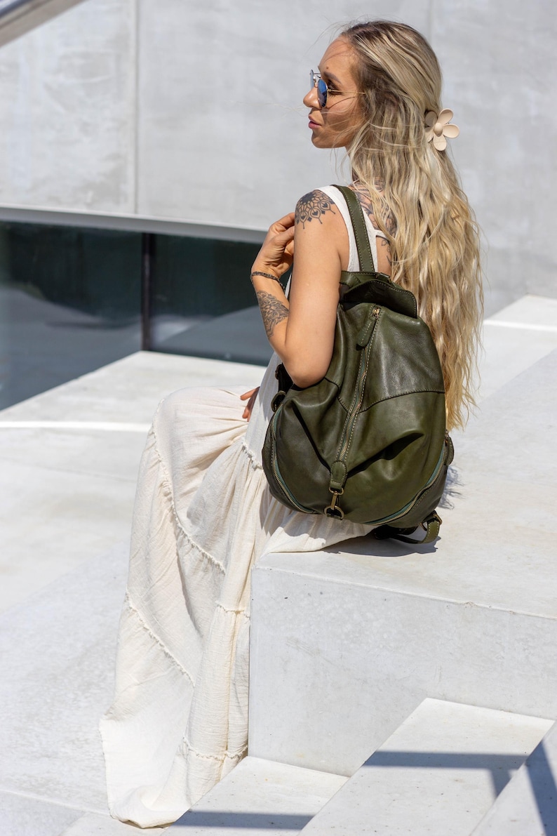 May include: An olive green leather backpack with a rounded silhouette, featuring a central vertical zip, a top flap closure, and adjustable shoulder straps. The bag has a smooth texture and antique brass-toned metal hardware accents.