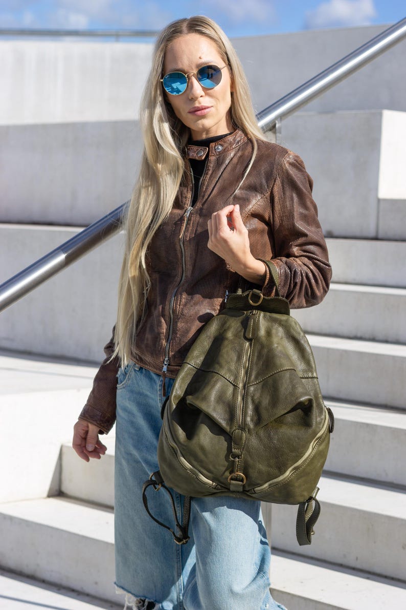 May include: An olive green leather backpack with a rounded shape, featuring a central vertical zip, a top handle, and adjustable shoulder straps. The bag has a distressed finish with antique brass hardware and a small buckle detail at the base.