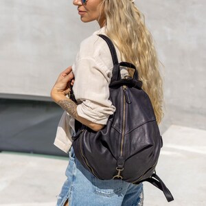 May include: A dark charcoal grey leather backpack featuring a rounded silhouette, a prominent vertical zippered front pocket, and adjustable shoulder straps. The bag includes antique brass-toned metal hardware, a top handle, and visible stitching.