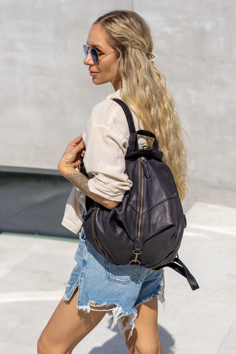 May include: A dark charcoal grey leather backpack featuring a rounded silhouette, a prominent vertical zippered front pocket, and adjustable shoulder straps. The bag includes antique brass-toned metal hardware, a top handle, and visible stitching.