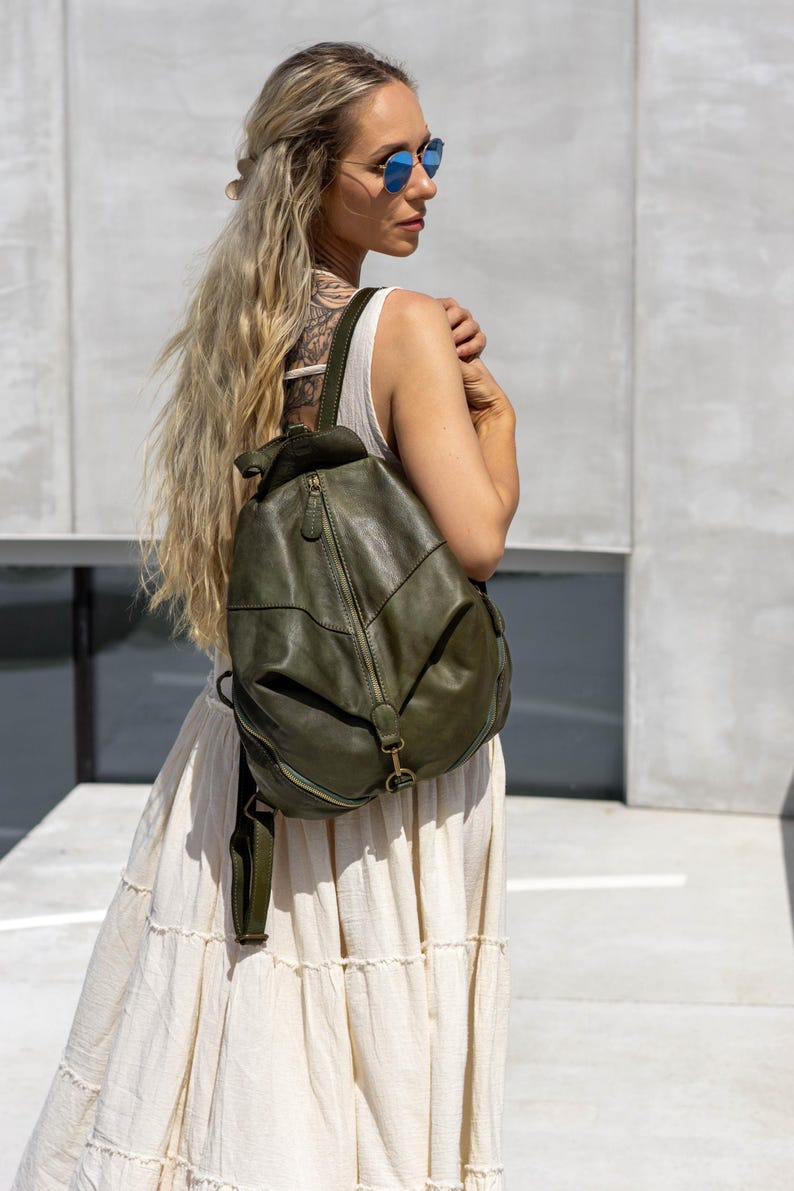 May include: An olive green leather backpack featuring a soft, slouchy silhouette, a vertical zippered front pocket, adjustable shoulder straps, and antique brass-toned metal hardware. The bag has a rounded shape with visible stitching details.