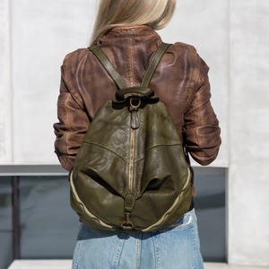 May include: An olive green leather backpack featuring a central vertical zip, a top handle, and adjustable shoulder straps. The bag has a rounded, slouchy silhouette with antique brass-toned metal hardware and a visible zip closure.