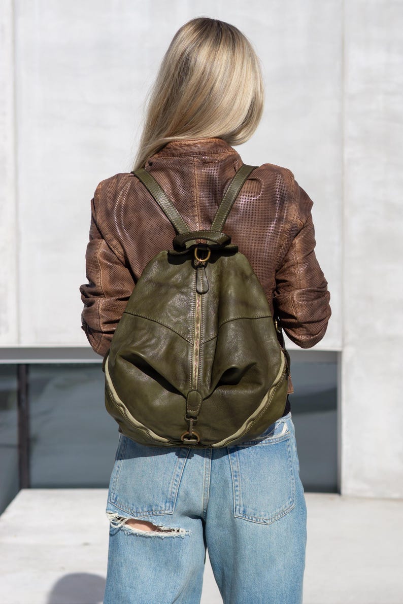 May include: An olive green leather backpack featuring a central vertical zip, a top handle, and adjustable shoulder straps. The bag has a rounded, slouchy silhouette with antique brass-toned metal hardware and a visible zip closure.