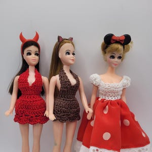 May include: Three fashion dolls are displayed, each with unique outfits and accessories. The first doll wears a red crocheted dress and devil horns. The second doll is in a brown crocheted dress and cat ears. The third doll has a red polka dot skirt, white top, and mouse ears.