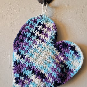 May include: A crocheted heart-shaped dishcloth in shades of blue, purple, and white, hanging from a hook on a white wall.