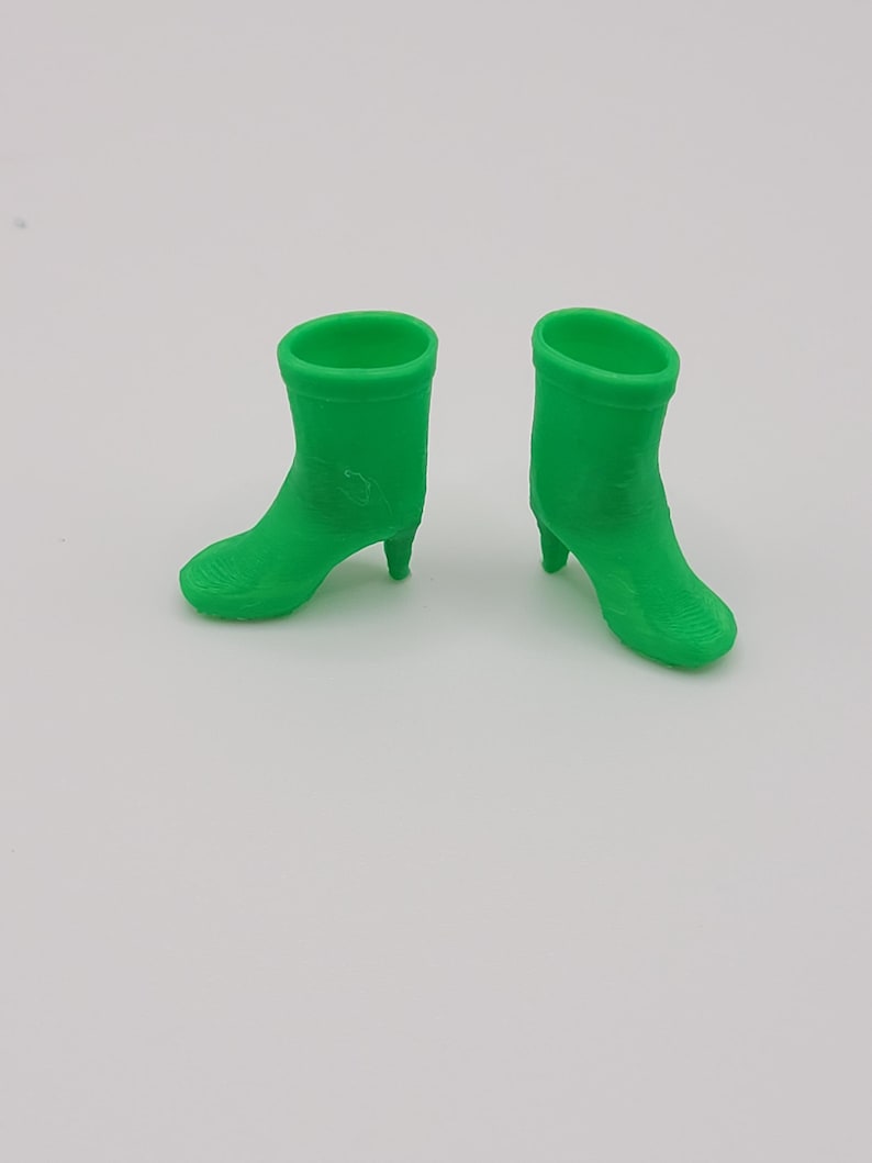 Puede incluir: Dos botines verdes brillantes con tacones peque&ntilde;os se muestran sobre un fondo blanco. Los botines tienen una textura suave y un dise&ntilde;o de punta redondeada. Los tacones son delgados y puntiagudos.