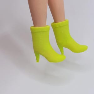 Puede incluir: Un par de botines verde ne&oacute;n con tacones de aguja. Los botines tienen una textura suave y una puntera redondeada. La imagen muestra los botines desde los tobillos hacia abajo, sobre un fondo blanco liso.
