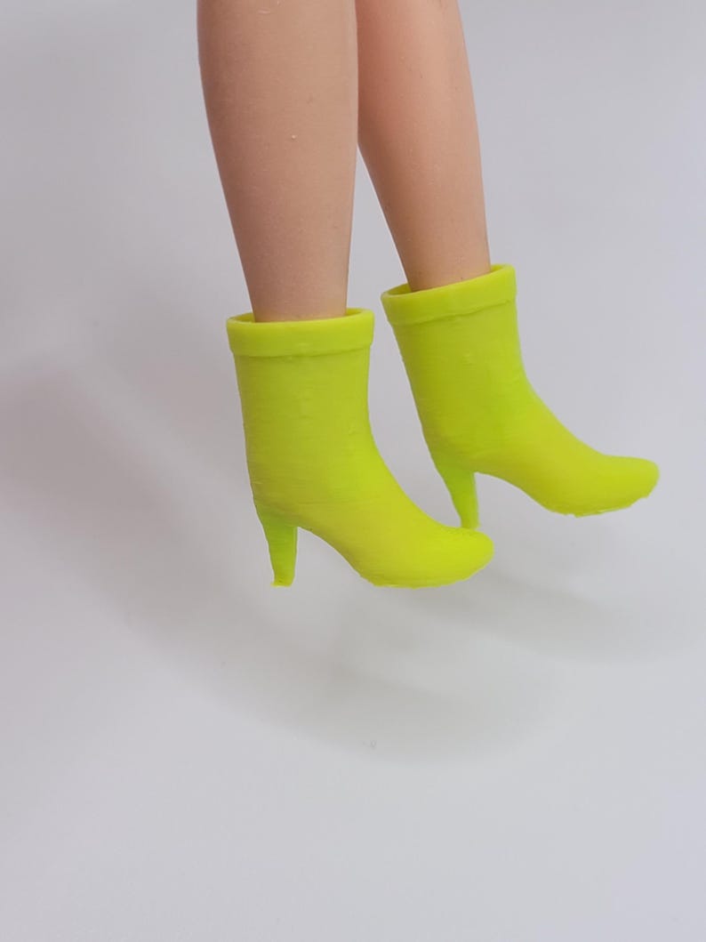 Puede incluir: Un par de botines verde ne&oacute;n con tacones de aguja. Los botines tienen una textura suave y una puntera redondeada. La imagen muestra los botines desde los tobillos hacia abajo, sobre un fondo blanco liso.
