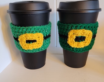 May include: Two black to-go coffee cups with green, yellow, and black crocheted cup cozies. The cozies feature a yellow buckle design.