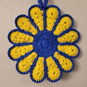 Può includere: Un sottopentola all'uncinetto a forma di fiore con filato blu e giallo. Il fiore ha 10 petali e un centro blu.