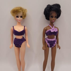 Pode incluir: Duas bonecas em pé lado a lado. A boneca à esquerda tem cabelo loiro e veste um biquíni de duas peças em crochê roxo escuro. A boneca à direita tem cabelo escuro e veste um biquíni de crochê roxo e lilás de duas peças com um laço.