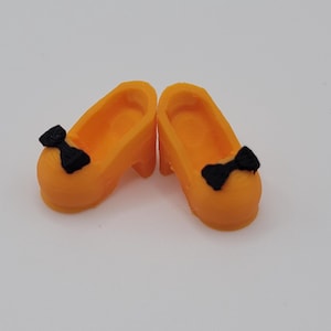 May include: A pair of orange doll shoes with small heels and black bow accents. The shoes are a bright, solid orange colour and appear to be made of plastic or a similar material. The bows are positioned on the front of each shoe.